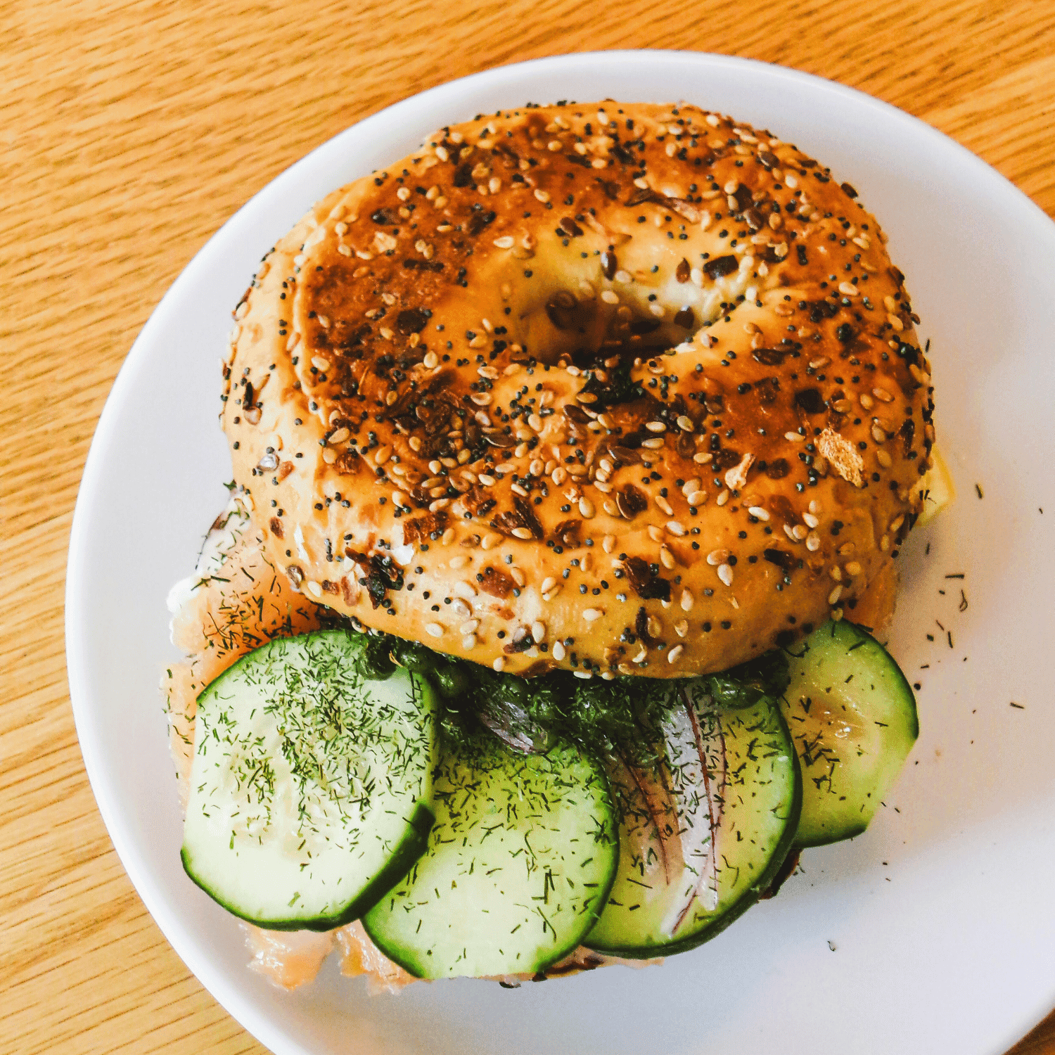 Bagel sandwich with cucumber, smoked salmon, cream cheese, and dill on a white plate.