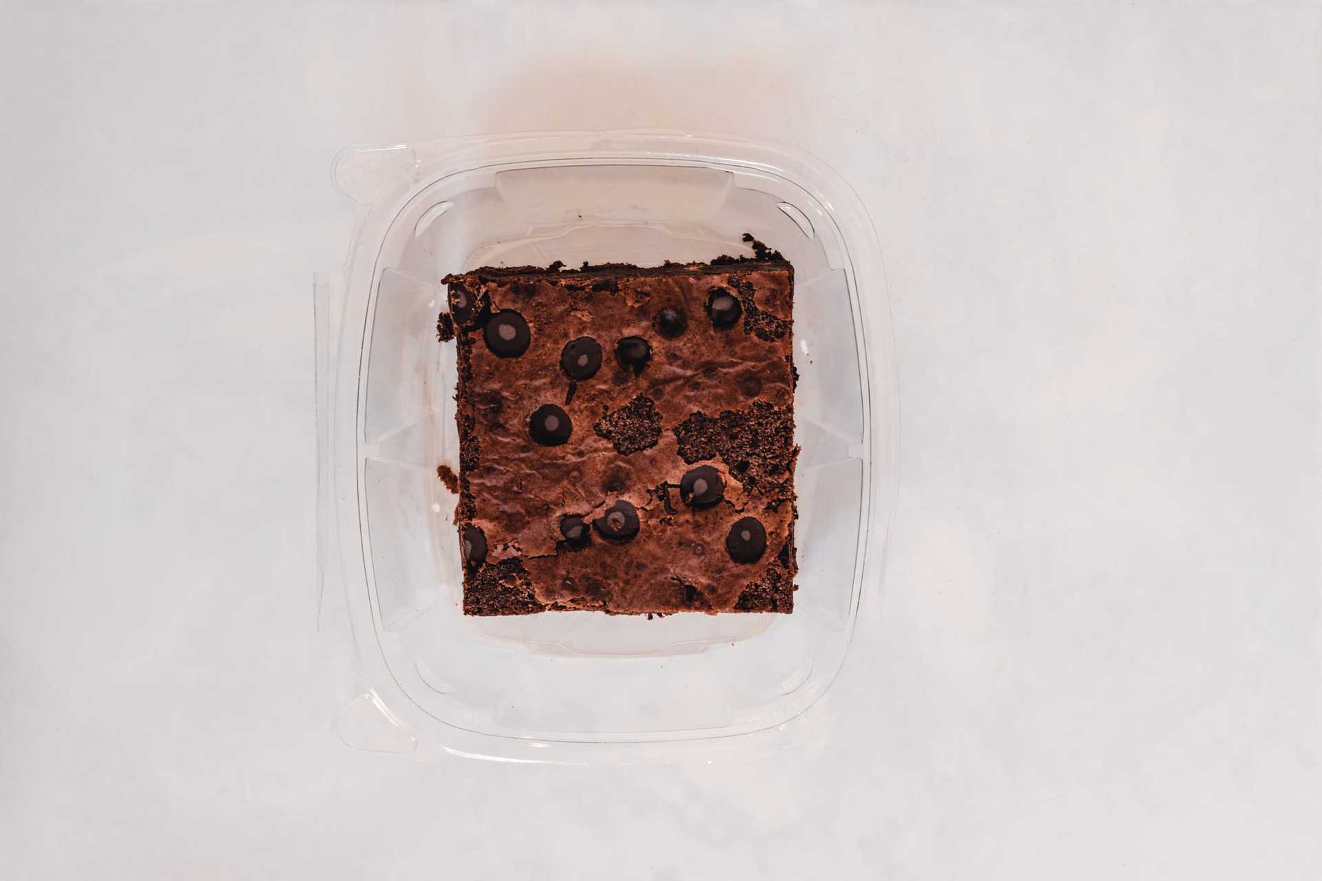 Chocolate brownie with chocolate chips in a clear plastic container on a white background.