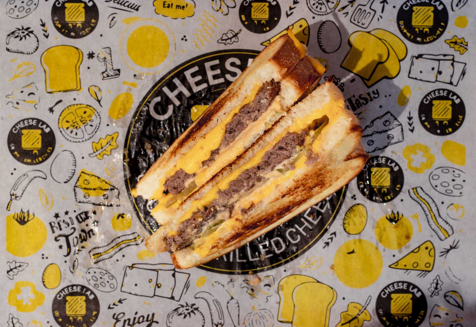 Grilled cheese sandwich with beef patty and cheddar on branded cheese-themed wrapping paper.
